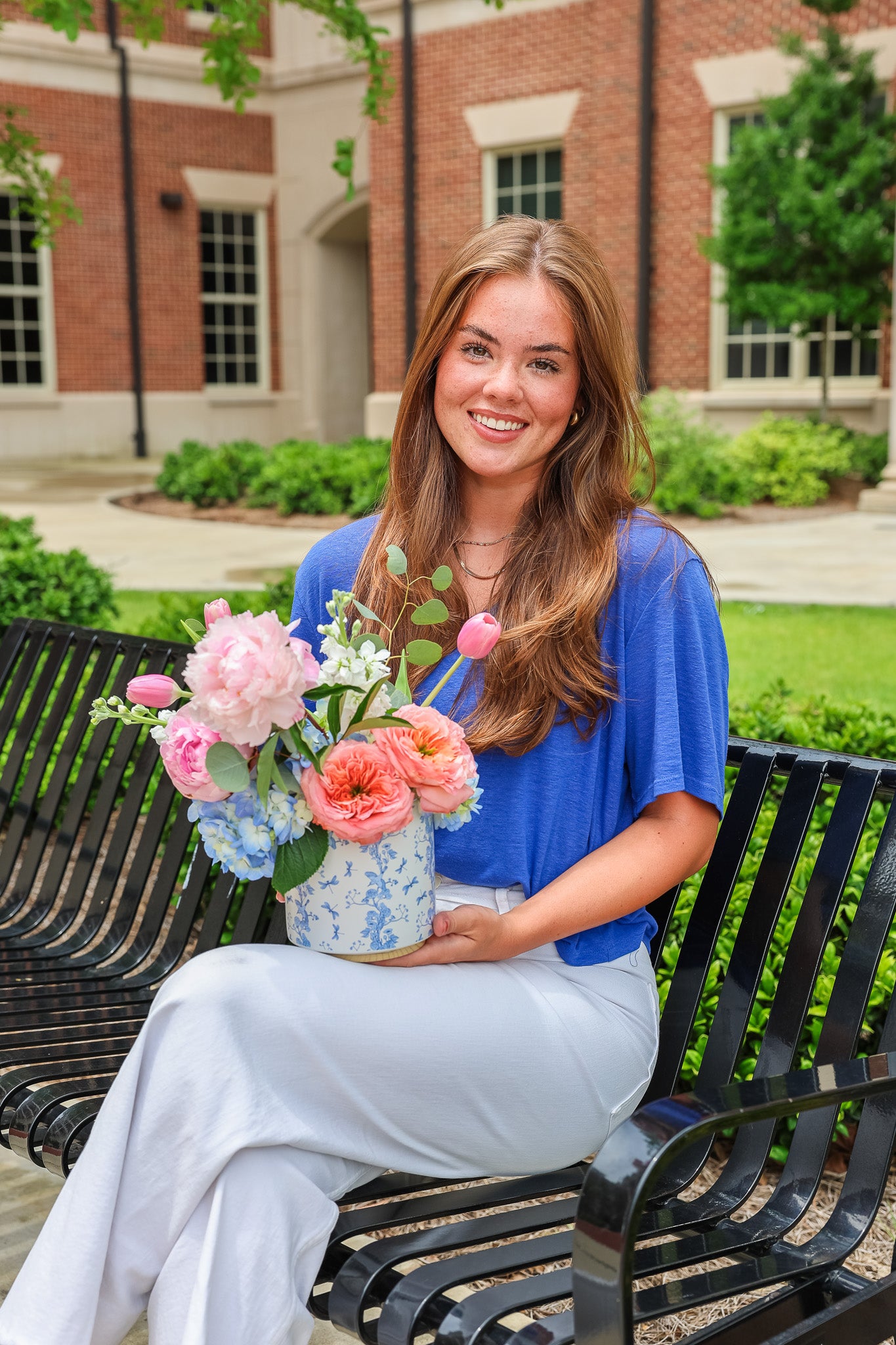 JJ sitting on a bench holding flowers outside of Tutwiler Hall on university of Alabama's campus